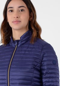 Giacca blu trapuntata con colletto alto e cerniera frontale. Presenta una superficie liscia e una toppa con logo sulla spalla sinistra.