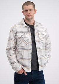 Witte geruite blouse met knopen, gemaakt van een gestructureerde stof met grijze, bruine en zwarte lijnen, voorzien van twee borstzakken en blauwe knopen.