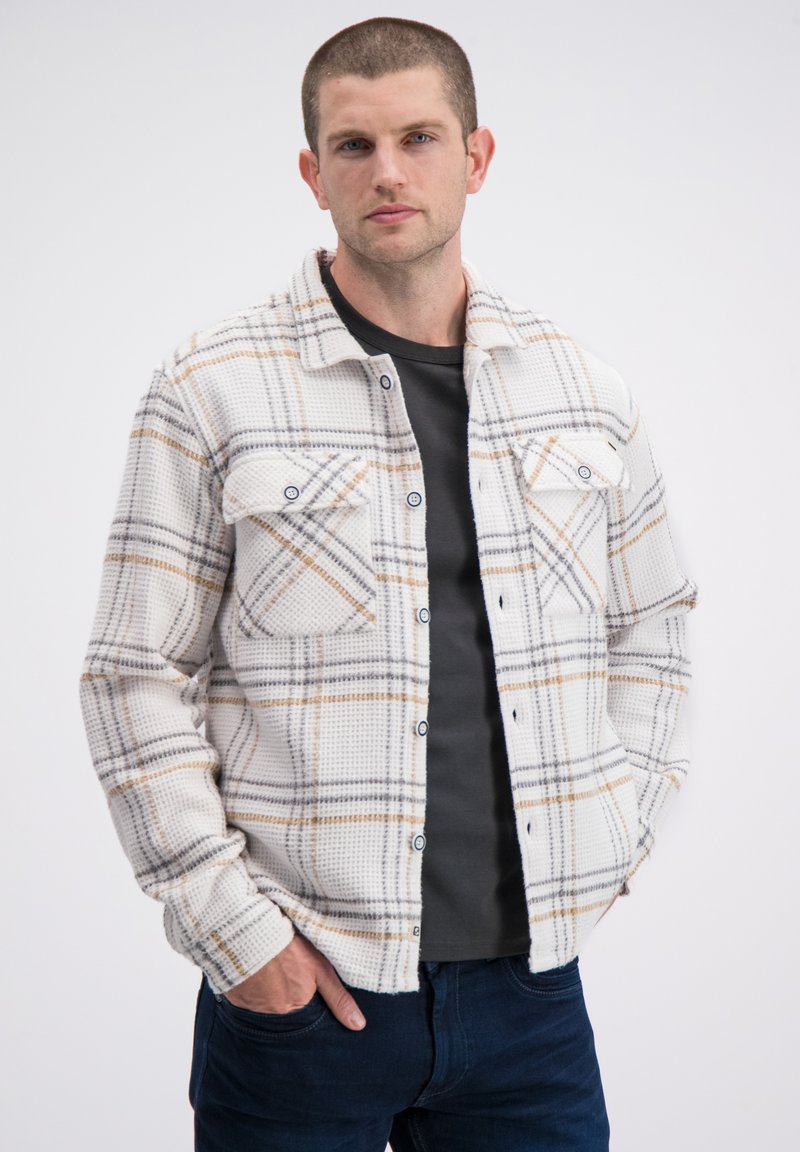 Witte geruite blouse met knopen, gemaakt van een gestructureerde stof met grijze, bruine en zwarte lijnen, voorzien van twee borstzakken en blauwe knopen.