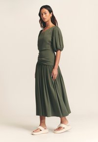 Abito verde oliva con maniche a sbuffo e gonna arricciata, realizzato in tessuto liscio. Abbinato a sandali con piattaforma di colore chiaro.