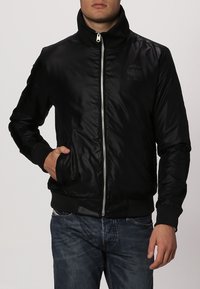 Svart bomberjacka med dragkedja framtill, hög krage, sidofickor och ribbade ärmslut. Glansigt yttre och subtil logotypdetalj på bröstet.