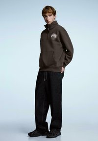 Brązowa bluza na zamek z przednią kieszenią, noszona z ciemnymi dżinsami. Na piersi znajduje się biały nadruk z tekstem. Całość uzupełniają czarne tenisówki.