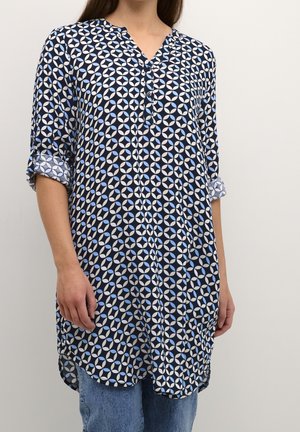 Kobieta ubrana w granatową tunikę z długim rękawem, ozdobioną białym i jasnoniebieskim wzorem w kółka, zapięta pod szyją na guziki, z podwiniętymi rękawami oraz jasnoniebieskie dżinsy.