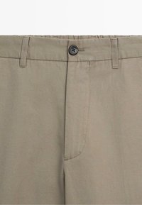 Pantalon beige avec fermeture à bouton, passants pour ceinture et détails visibles sur la couture avant.
