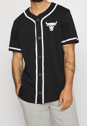 Mann trägt schwarzes, geknöpftes Baseballhemd mit weißem Saum und Stierkopf-Logo auf der Brust, hellgraue Hose und sichtbare Arm-Tattoos.
