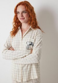 Cremfarbendes karierter Button-Up-Hemd mit langen Ärmeln, einer Brusttasche mit einem gestickten Cartoon-Charakter und einem klassischen Kragendesign.