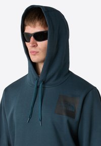 Donkerblauwe hoodie met een zwart logo op de zak, voorzien van een voorzak en verstelbare koorden. Draagt een paar donkere zonnebrillen.