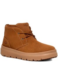 Botas de ante marrón hasta el tobillo con suelas de goma planas, detalles de cordones y un diseño minimalista. Logotipo de UGG en el lateral y en la parte superior.