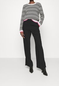 Schwarze weit geschnittene Hosen mit seitlichen Schlitzen, kombiniert mit einem grau-schwarz gemusterten Pullover, der rosa Akzente an den Bündchen und dem Saum hat.