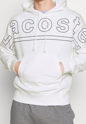 Person trägt einen weißen Hoodie mit großen schwarz umrandeten Buchstaben auf Brust und Schultern, die Hände in der Fronttasche, kombiniert mit grauer Hose.