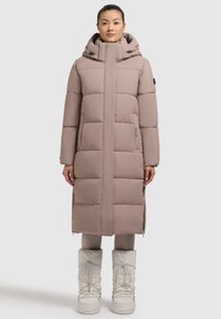 Palton lung căptușit, de culoare bej deschis, cu guler înalt și glugă. Prezintă secțiuni matlasate orizontale și buzunare frontale. Purtați cu cizme albe.