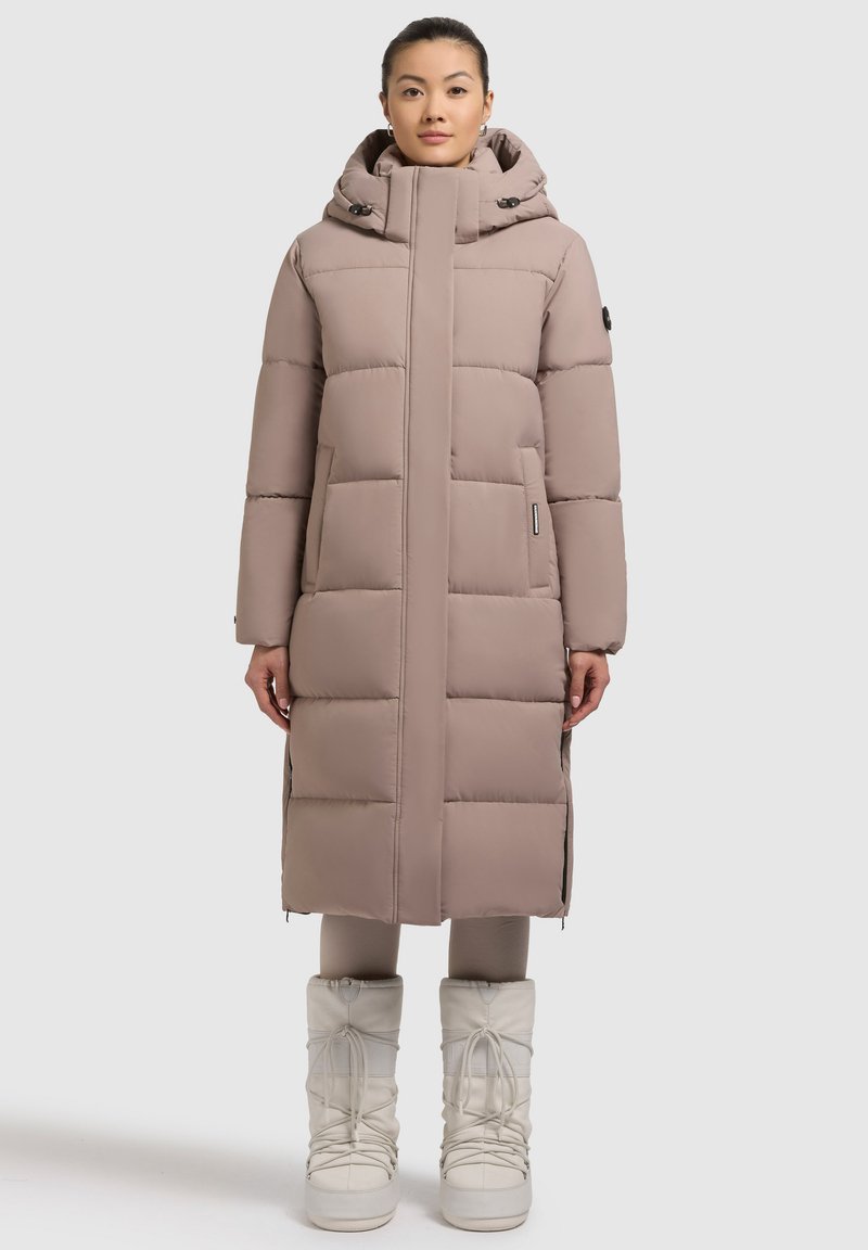 Palton lung căptușit, de culoare bej deschis, cu guler înalt și glugă. Prezintă secțiuni matlasate orizontale și buzunare frontale. Purtați cu cizme albe.