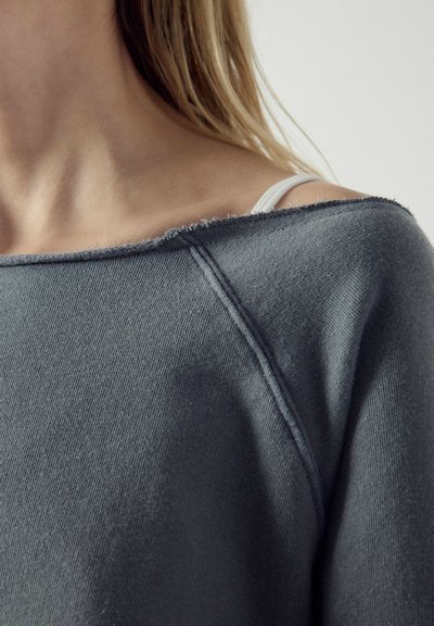 Gros plan sur l’épaule d’une personne portant un pull gris ample avec un ourlet brut, révélant une bretelle de soutien-gorge blanche en dessous.