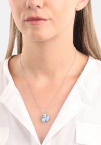 Srebrny naszyjnik łańcuszkowy z dużym sercowym zawieszką w jasnoniebieskim kolorze oraz małym kwiatowym akcentem na górze.