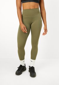 Person som bär olivgrön högmidjebyxor, svart sport-bh, vita Nike-strumpor och svarta sneakers, står mot en vit bakgrund.