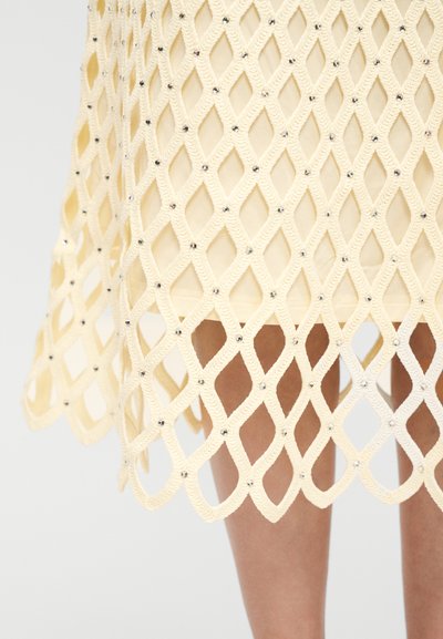Gros plan sur une jupe en filet tissé crème avec de petites perles argentées, laissant entrevoir les jambes à travers un motif ajouré en forme de losange.