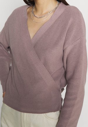 Frau trägt einen mauvefarbenen Strick-Wickelpullover mit V-Ausschnitt und geschichteten silbernen Ketten, kombiniert mit hellbeigen Hosen.