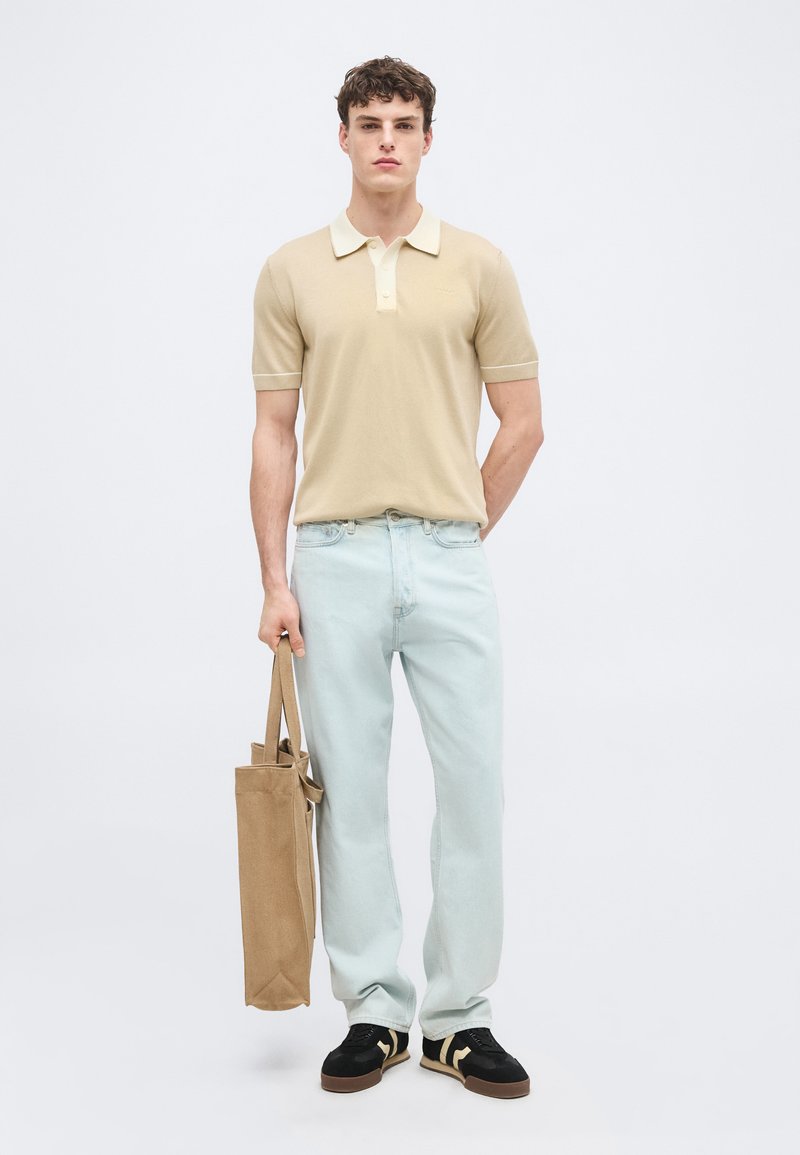 Jeune homme portant un polo beige, un jean bleu clair, des baskets noires, tenant un sac fourre-tout beige, debout devant un fond blanc uni.