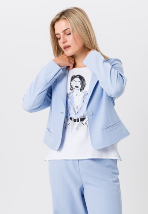 Blazer ajusté bleu clair avec une texture lisse, deux poches avant et fermeture à boutons, porté sur un t-shirt blanc graphique avec une illustration noire.