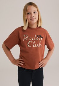 Rūsas krāsas kokvilnas t-krekls ar īsām piedurknēm, kurā ir iestrādāti rakstaini izšūti burti "Rodeo Club" un neliela melna kovboja cepure kā akcents.