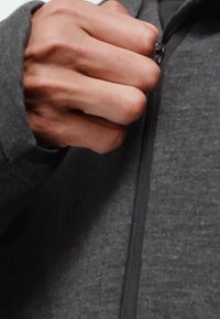 Hand gripping zipper of dark gray zip-up jacket, pulling it upward against a plain background.