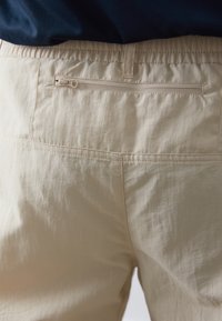 Beige kalhoty s elastickým pasem a kapsou se zipem na zadní straně. Materiál je lehký a mírně texturovaný, se šitými detaily švů.