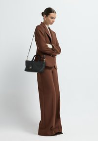 Completo marrone su misura con pantaloni a gamba larga e blazer aderente. Caratterizzato da bottoni dorati. Borsa nera intrecciata con manici corti e tracolla.