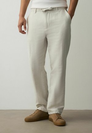 Person trägt hellbeige Leinenhose mit seitlichen Taschen und beigefarbenen Schnürschuhen, steht auf einem grauen Boden vor einer schlichten Wand.