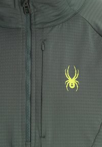 Veste zippée verte en tissu texturé, avec un logo d'araignée jaune bien en vue et une fermeture éclair à l'avant.