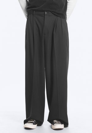 Homme portant un pantalon noir à larges jambes à fines rayures et des baskets noires et blanches, debout devant un fond blanc.
