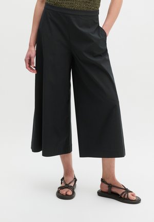 Femme portant un pantalon noir large et court et des sandales plates noires à brides, debout avec une main dans la poche sur fond blanc.
