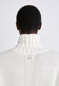 Weißer Strickpullover mit hohem, geripptem Kragen, strukturierter Webart und einem kleinen metallischen Etikett im Nackenbereich.