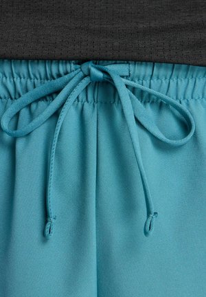 Primo piano della vita elastica turchese con coulisse annodata su pantaloni leggeri, abbinata a tessuto nero testurizzato sopra.