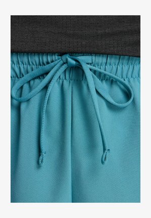 Close-up of teal elastic waistband with tied drawstring on lightweight pants, paired with black textured fabric above.