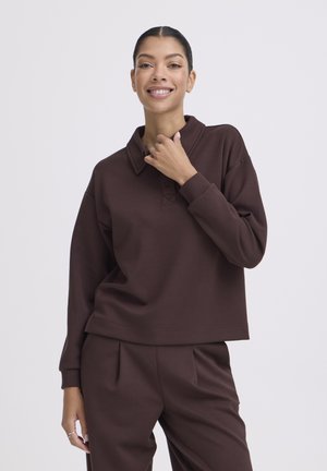 Mujer sonriente con cabello oscuro en un moño bajo, vistiendo una sudadera suelta de cuello y color marrón oscuro y pantalones plisados a juego sobre un fondo liso.