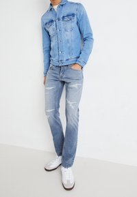 Ljusblå jeansjacka med två bröstfickor, tillsammans med ljusblå slitna jeans och vita sneakers med röda detaljer.