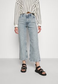 Ljust blå, hög midja, vida jeans med rå nederkant. Matchas med en croppad, krämfärgad och svart randig tröja och svarta sandaler.