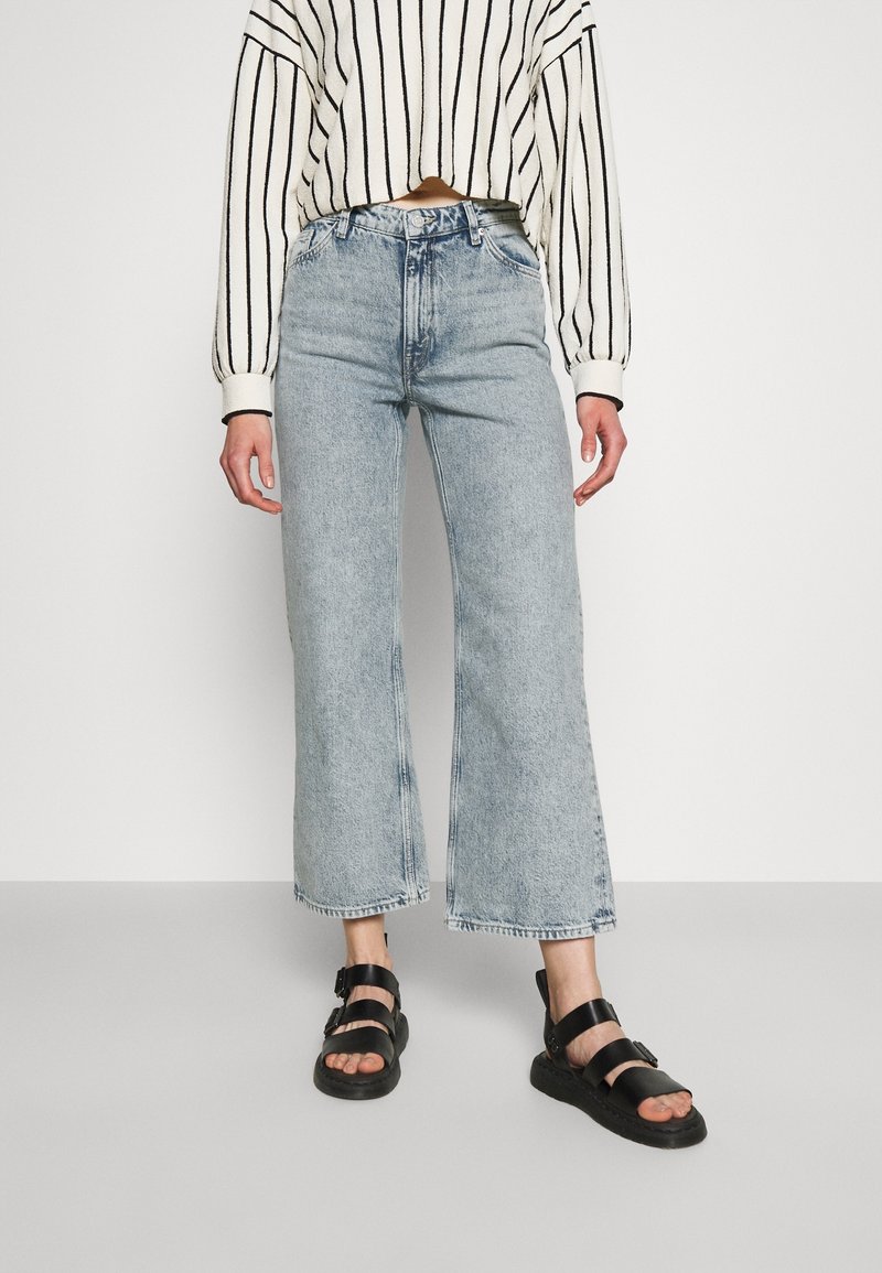 Ljust blå, hög midja, vida jeans med rå nederkant. Matchas med en croppad, krämfärgad och svart randig tröja och svarta sandaler.