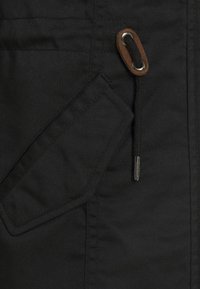 Detalles de chaqueta negra que incluyen un bolsillo diagonal, ojales de metal y un cordón con un acento de cuero marrón. Tejido texturizado, diseño estructurado.