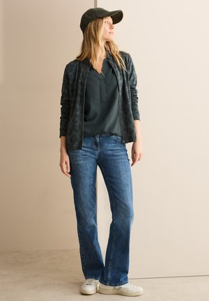 Veste zippée vert foncé avec imprimé léopard, blouse vert foncé, jeans évasés bleus et baskets blanches, le tout complété par une casquette verte.