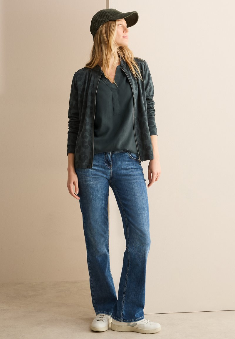 Donkergroene zip-up jas met een luipaardprint, donkergroene blouse, blauwe flared jeans en witte sneakers, afgewerkt met een groene pet.