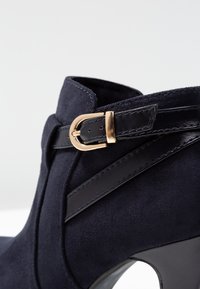 Botín de tobillo en color navy con textura de ante, una silueta elegante y un detalle de hebilla dorada en la correa del tobillo.