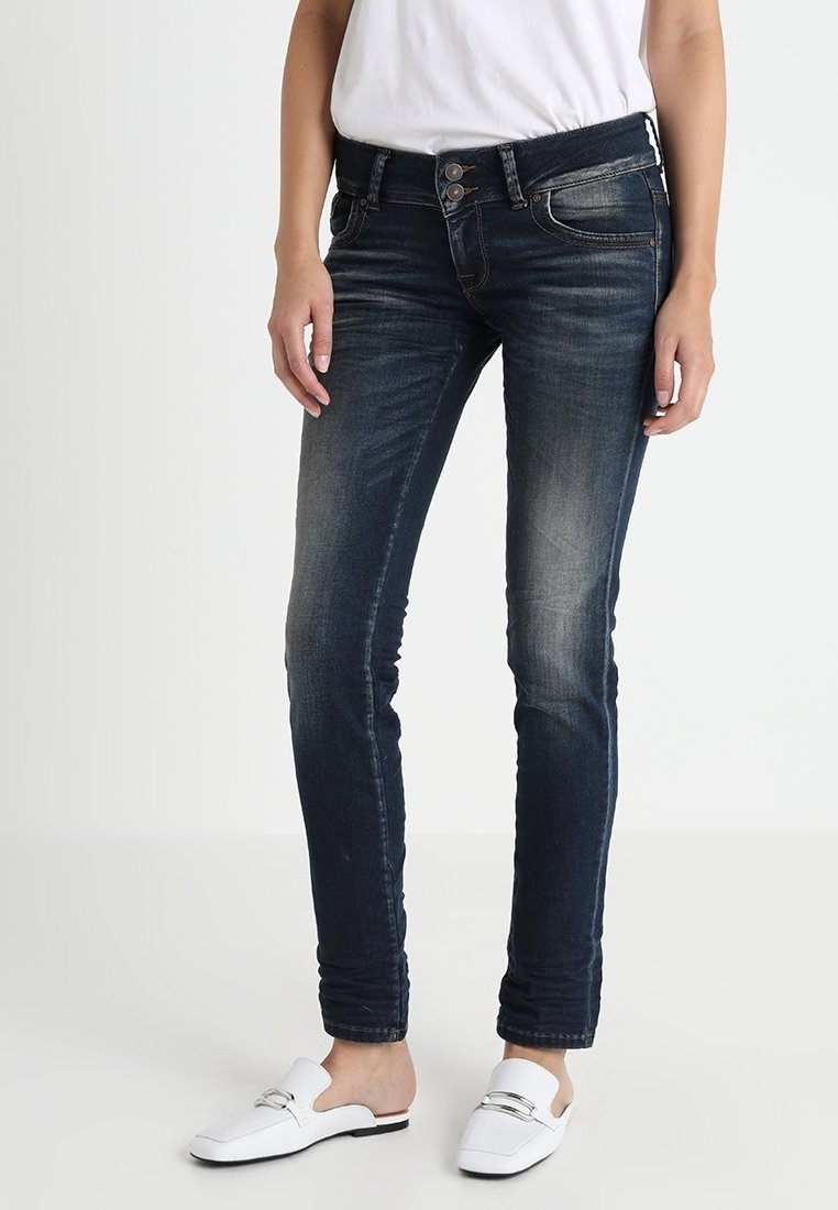 Femme portant un jean slim bleu foncé avec des détails délavés, une chemise blanche et des mocassins blancs à enfiler avec des accents de boucle argentés.