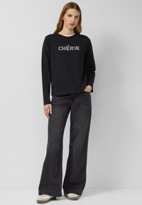 Czarna bluzka z długim rękawem z białym napisem "CHÉRIE", zestawiona z szerokimi ciemnymi jeansami i beżowymi trampkami. Prosty, casualowy zestaw.