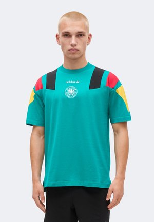 Junger Mann trägt ein türkisfarbenes Adidas T-Shirt mit schwarzen, roten und gelben Aufnähern und deutschem Fußball-Emblem, steht vor einem einfarbigen hellen Hintergrund.