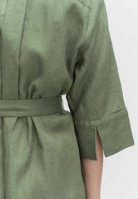 Zielona lniana koszula z paskiem w talii, z podwiniętymi rękawami i bocznymi szlicami, eksponująca gładką fakturę i swobodny styl.