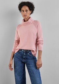 Hellrosa Rollkragensweater mit Knopfdetails und strukturierten Akzenten an den Schultern, kombiniert mit blauen weit geschnittenen Jeans. Hauchweiche Strickware.