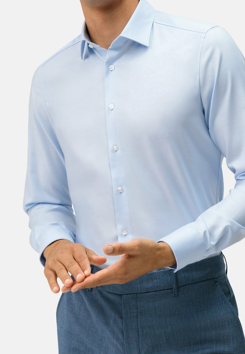 Chemise bleu clair boutonnée avec une texture lisse, dotée d'un col pointu et de manches longues, portée avec un pantalon bleu foncé.