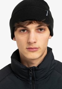 Berretto a coste nera con un piccolo tag logo, indossato con un giubbotto nero a colletto alto. Primo piano di un volto con espressione neutra.