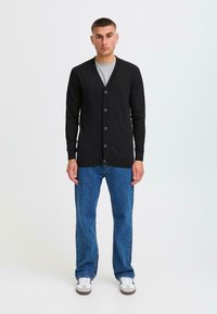 Schwarze Strickjacke mit V-Ausschnitt, ausgestattet mit sechs runden Knöpfen, kombiniert mit blauen Jeans und weißen Sneakers, vor einem schlichten Hintergrund stehend.